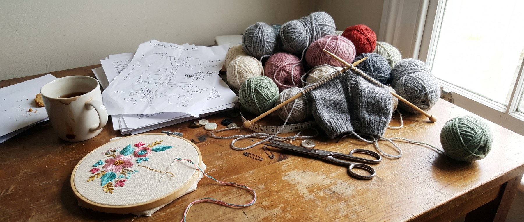 A corner of Margaret's desk: an embroidery hoop with a half-finished spray of flowers, balls of merino in grey, cream and blush, brass scissors, buttons and a cardigan on the needles